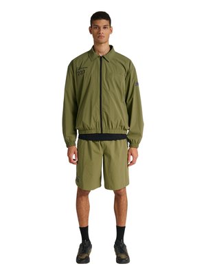 Junger Mann steht, trägt eine olivgrüne Jacke mit Reißverschluss und passende Shorts, schwarze Socken und dunkle Sneakers, blickt nach vorne auf weißem Hintergrund.