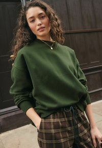 Sudadera verde oliva con cuello redondo y hombros caídos, combinada con pantalones de cuadros marrones con cordón en la cintura que presentan un tejido texturizado.