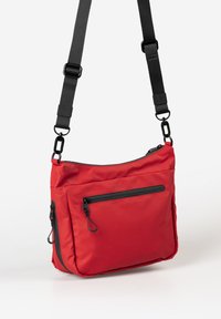 Rote Nylon-Schultertasche mit verstellbarem schwarzen Tragegurt, ausgestattet mit einer vorderen Reißverschlusstasche und zwei seitlichen Reißverschlussfächern. Glatte Textur.