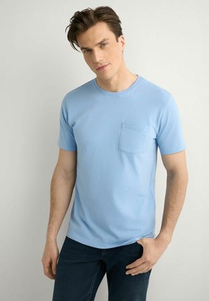 Mężczyzna ubrany w jasnoniebieską koszulkę z krótkim rękawem i kieszenią na piersi, ciemne dżinsy, stoi na tle jednolitego jasnego tła, spogląda w kamerę.