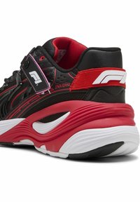 Chaussure de sport noire et rouge avec une tige en mesh texturé, une semelle rouge amortie, des lignes de design audacieuses et une sangle ajustable arborant un logo.