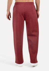 Bordeauxrøde joggingbukser lavet af blødt, strækbart materiale med løs pasform, elastisk talje og sidelommer. Hvide sneakers ses nederst.