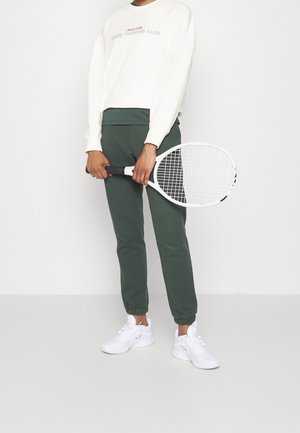 Persona che indossa una felpa bianca e pantaloni sportivi verdi, tiene una racchetta da tennis bianca con entrambe le mani di fronte al corpo.