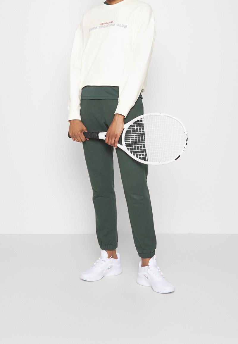 Persona che indossa una felpa bianca e pantaloni sportivi verdi, tiene una racchetta da tennis bianca con entrambe le mani di fronte al corpo.