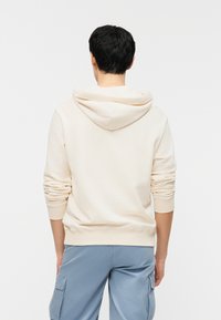 Άτομο που φοράει μια μπεζ φούτερ με κουκούλα κατεβασμένη και ανοιχτό μπλε παντελόνι cargo, στέκεται με την πλάτη προς το μέρος μας σε ένα απλό λευκό φόντο.