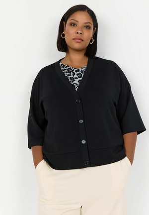 Černý oversized cardigan s krátkými rukávy, výstřihem do V a šesti knoflíky. Přes světlomodrý top s leopardím vzorem. Hladká textura.