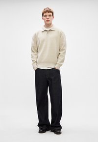 Pulover polo tricotat bej cu guler, asortat cu jeansi evazați de culoare albastru închis. Pantofi negri vizibili. Modelul stă �în fața unui fundal simplu.