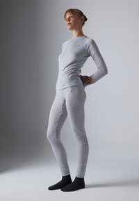 Una donna è in piedi con le mani dietro la schiena, indossando un top aderente grigio chiaro a maniche lunghe e leggings, e calze nere, su uno sfondo semplice.