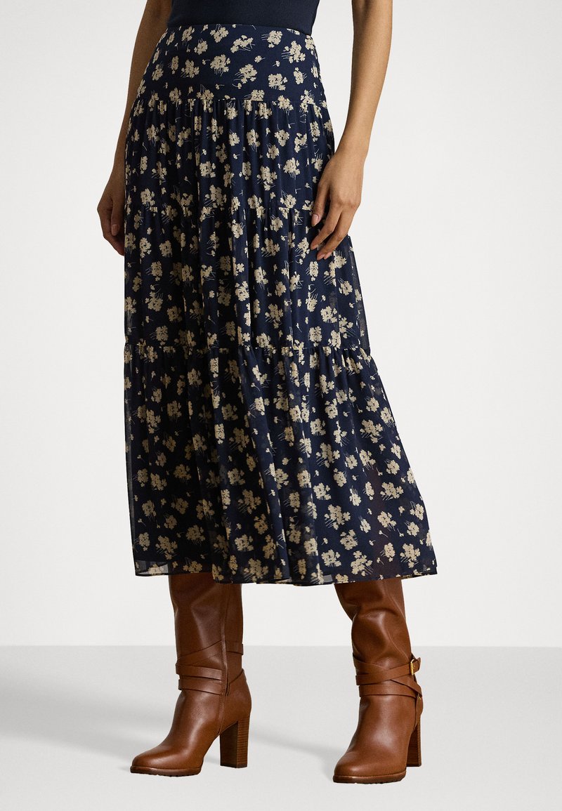 Gonna maxi floreale blu navy con design a strati; presenta piccoli fiori beige su un tessuto fluente, abbinata a stivali alti fino al ginocchio marroni con tacco.