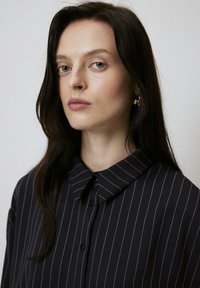 Navy button-up shirt with white vertical stripes, featuring a pointed collar and smooth texture. Hair styled down, pendant earrings are visible.