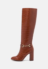 Botte haute jusqu'au genou en cuir marron embossé crocodile, avec bout pointu et talon épais. Dotée d'accents en métal doré et d'une coupe ajustée.