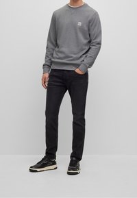 Felpa grigia girocollo con polsini e orlo a coste, abbinata a jeans neri slim fit e sneakers nere con dettagli bianchi.