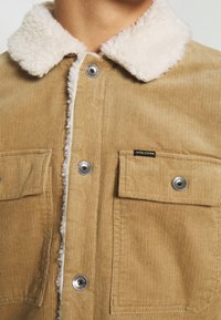 Beżowa kurtka z korduroju z kremowym kołnierzem z sztucznego barana, z dwiema kieszeniami na piersi, srebrnymi zatrzaskami i teksturowanymi pionowymi paskami.