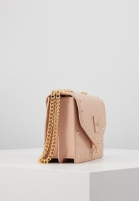 Malá kabelka z růžové kůže s zlatými hřebíky a zlatým řetízkovým popruhem na bílém podstavci na šedém pozadí.