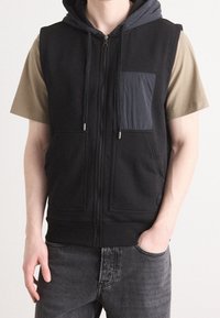 Muž v černé bezrukávové zipové mikině s předními kapsami, pod kterou má béžové tričko s krátkým rukávem, a tmavě šedé džíny, s jednou rukou v kapse.