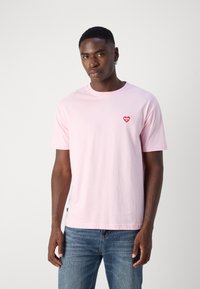 Lichtroze katoenen T-shirt met een ronde hals, korte mouwen en een kleine rode hartafbeelding op de linkerkant van de borst.