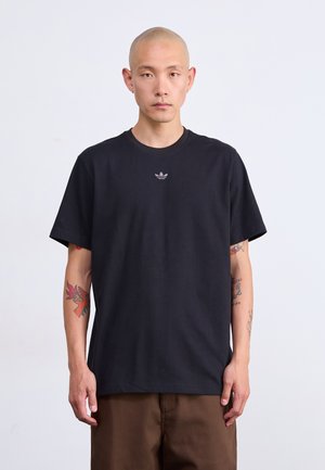 Borotvált fejű férfi fekete Adidas pólóban és barna nadrágban, színes tetoválásokkal mindkét alkarján, sima háttér előtt áll.