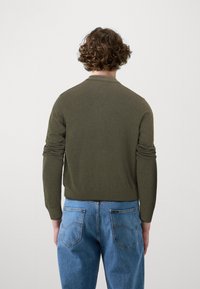 Suéter de punto verde oliva con mangas largas y puños acanalados, combinado con jeans de mezclilla azul claro que muestran las costuras de los bolsillos traseros.