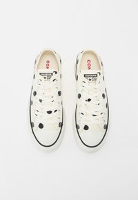 Platnene tenisice u krem boji s crnim točkicama, bijelim vezicama i čepom za prste. Imaju istaknuto "CONVERSE" brendiranje na jeziku i sa strane.