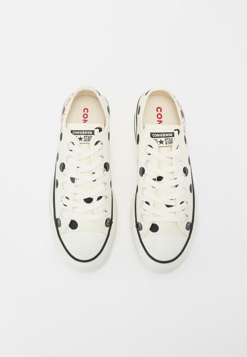 Zapatillas de lona en color crema con lunares negros, cordones blancos y punta reforzada. Presentan un llamativo logotipo de "CONVERSE" en la lengüeta y en el lateral.
