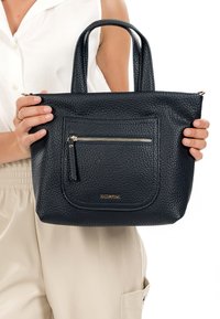 Zwarte tas van geruwd leer met dubbele handvatten, een voorvak met rits en subtiele gouden accenten. Compacte, rechthoekige vorm.