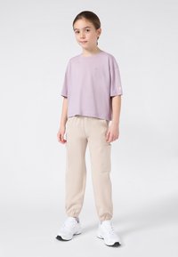 Jeune personne debout, portant un t-shirt oversize lavande, un pantalon cargo beige, des baskets blanches, et regardant légèrement sur le côté.