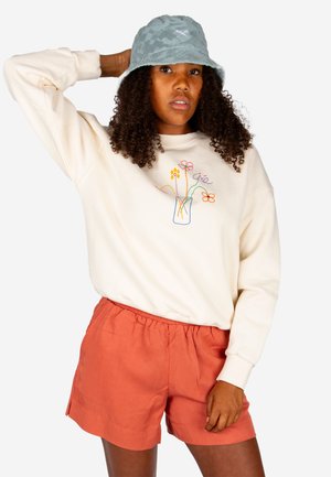 Woman with curly hair wearing a green bucket hat, cream sweatshirt with colorful flower embroidery, and orange shorts, posing with hand behind head.