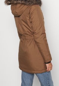 Brown insulated jacket with a faux fur hood, cinched waist, and long sleeves, paired with light blue jeans. Smooth texture, functional design.