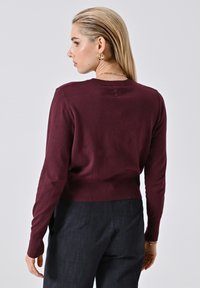Frau mit slicked-back blonden Haaren trägt goldene Hoop-Ohrringe, einen burgunderfarbenen Pullover und dunkle Hose, steht vor einem schlichten hellen Hintergrund.