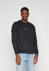 Felpa nera con scollatura a girocollo, polsini e orlo a costine. Presenta un piccolo logo bianco e rosso sul petto. Indossata con jeans azzurri chiari.