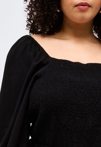 Γυναίκα με κατσαρά μαλλιά που φοράει μια μαύρη μπλούζα χωρίς ώμους με ελαστική υφή και μακριά φουσκωτά μανίκια, σε απλό φόντο.