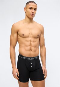 Μυώδης άνδρας που φορά μαύρα μποξέρ Tommy Hilfiger, στέκεται μπροστά σε απλό φωτεινό φόντο, κοιτάζοντας την κάμερα με ουδέτερη έκφραση.