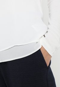 Persona con una camicia bianca a maniche lunghe con orlo a strati e pantaloni blu scuro con piccoli punti bianchi, con una mano appoggiata in tasca.