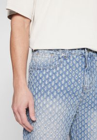 Närbild av en persons vänstra hand som vilar vid sidan, bärande ljusblå slitna denimjeans och en kortärmad beige skjorta.