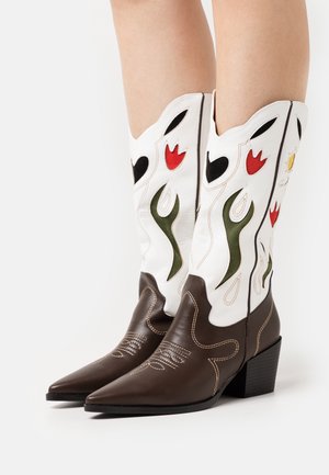 Hnědé kožené kovbojské boty s špičatou špičkou a středním podpatkem, s bílými návleky vyšívanými červenými, zelenými a černými květinovými vzory.