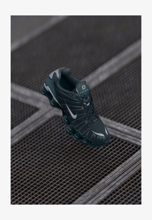 Zapatilla Nike Shox de color verde oscuro con parte superior de malla, acentos de goma y suela acolchada distintiva. Logo de Nike en plata en el lateral. Detalles texturizados visibles.