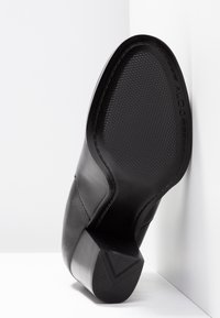 Zapato de tacón negro de cuero con una suela texturizada y forma de bloque geométrico. Acabado suave, con una marca discreta en la suela.