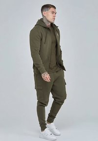 Olivgrüner Hoodie- und Jogginganzug aus weichem Stoff. Verfügt über einen Reißverschluss, Seitentaschen, elastische Bündchen und weiße Sneakers.