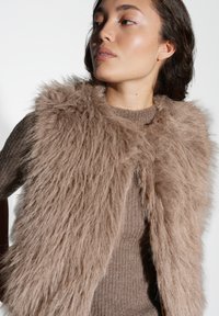 Eine Frau trägt einen beigen, gerippten Pullover und eine flauschige, hellbraune Fellweste. Sie blickt zur Seite vor einem schlichten weißen Hintergrund.