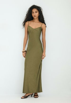 Femme aux longs cheveux bouclés portant une robe sans manches vert olive à fines bretelles et des sandales noires à talons hauts, debout devant un fond clair uni.