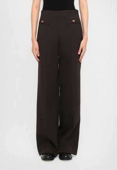 Femme portant un pantalon large taille haute marron foncé avec des détails de boutons dorés et des chaussures noires, se tenant devant un fond uni.