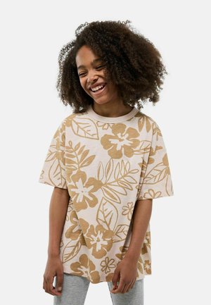 T-shirt oversize beige clair avec un imprimé floral et de feuilles marron. Manches courtes, encolure ras du cou et texture douce. Assorti à un pantalon gris.