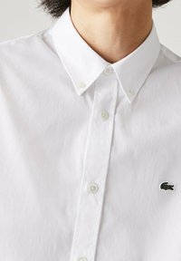 White collared shirt made of cotton with a smooth texture, featuring a small green embroidered crocodile logo and white buttons.