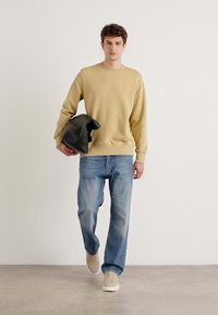 Lichtbeige sweatshirt, blauwe denim jeans en beige sneakers. Model met een zwarte tas, staand op een grijze vloer met een egale achtergrond.