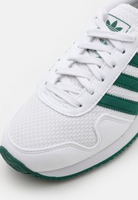 Vit sportskor med gröna mockadetaljer och tre ränder. Mönstrad mesh-överdel med sn örning och gummisulor.