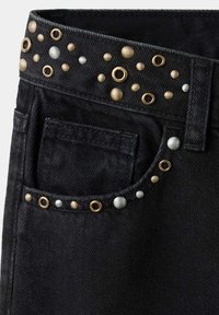 Poche et taille d’un jean en denim noir décorées de petits clous ronds métalliques dorés et argentés.