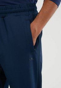 Marineblauwe sportbroek gemaakt van gladde stof, met een elastische tailleband, zijzakken en een klein merksymbool op de dij.