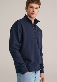 Tengerészkék cipzáras pulóver magas gallérral, bordázott mandzsettákkal és szegéllyel. Kis hímzett logó található a mellkason. Puha szövet textúra.