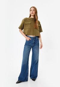 Olijfgroen cropped top met twee voorzakken, gecombineerd met wijde blauwe denim jeans en zwarte puntige schoenen. Eenvoudig ontwerp en ontspannen pasvorm.