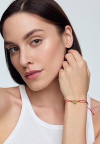 Złota bransoletka z zawieszką w kształcie serca na czerwonym sznurku. Sznurek jest związany małymi metalowymi akcentami z obu stron, noszona na nadgarstku na jasnym tle.